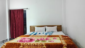 A bed with a floral bedspread, two pillows, and a wooden headboard in a room with white walls and a red curtain. - Homecation Manas River Rock Lodge (Barnagar)