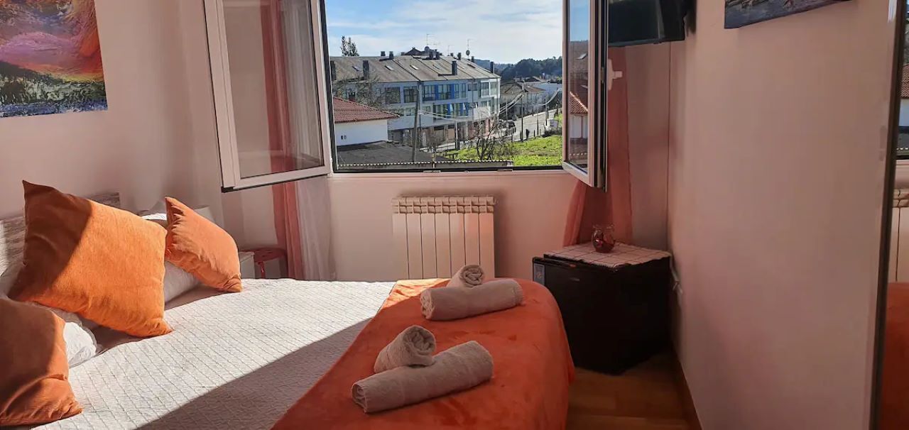 Une chambre à coucher avec un lit, des coussins orange, une fenêtre donnant sur des maisons et un radiateur.