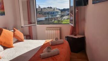 Une chambre à coucher avec un lit, des coussins orange, une fenêtre donnant sur des maisons et un radiateur.