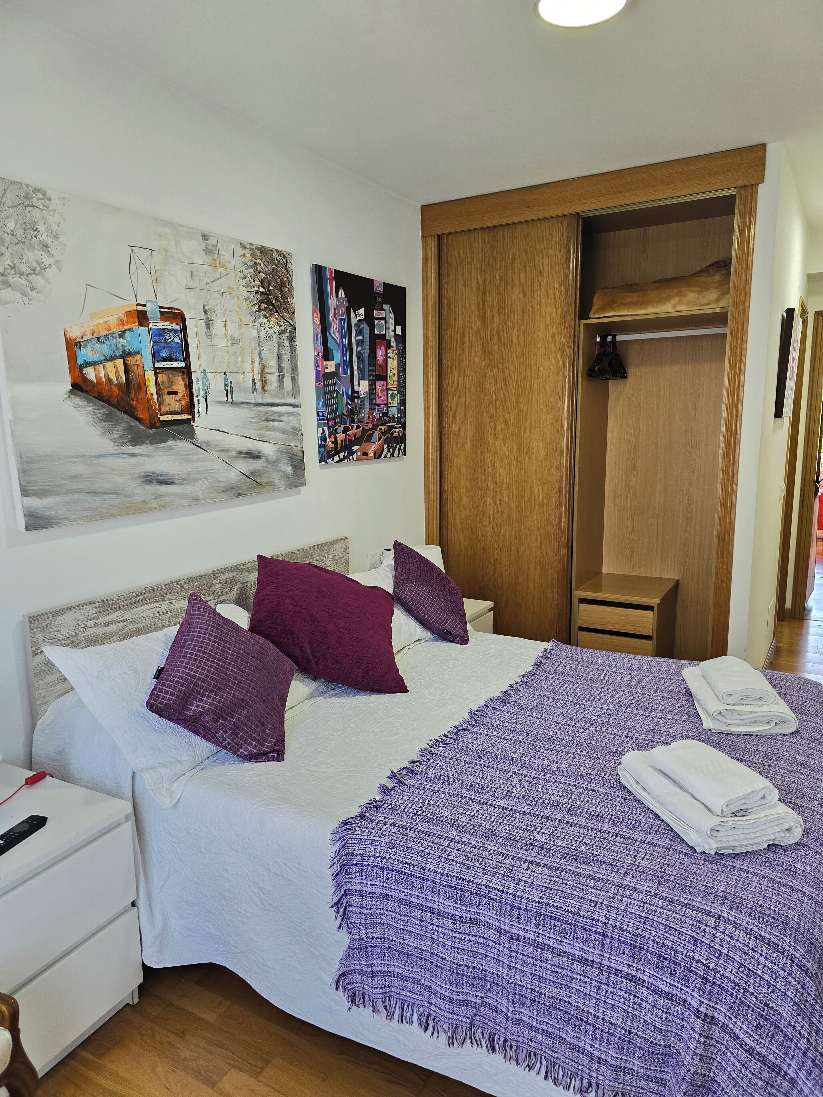 Un lit bien fait, recouvert d’une couverture violette, une armoire en bois et un tableau représentant un paysage urbain accroché au mur.