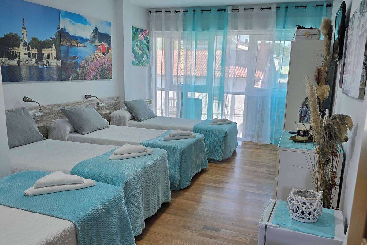 A hotel room with four beds arranged in a row, a wooden floor, a desk with a chair, and a large window with curtains.