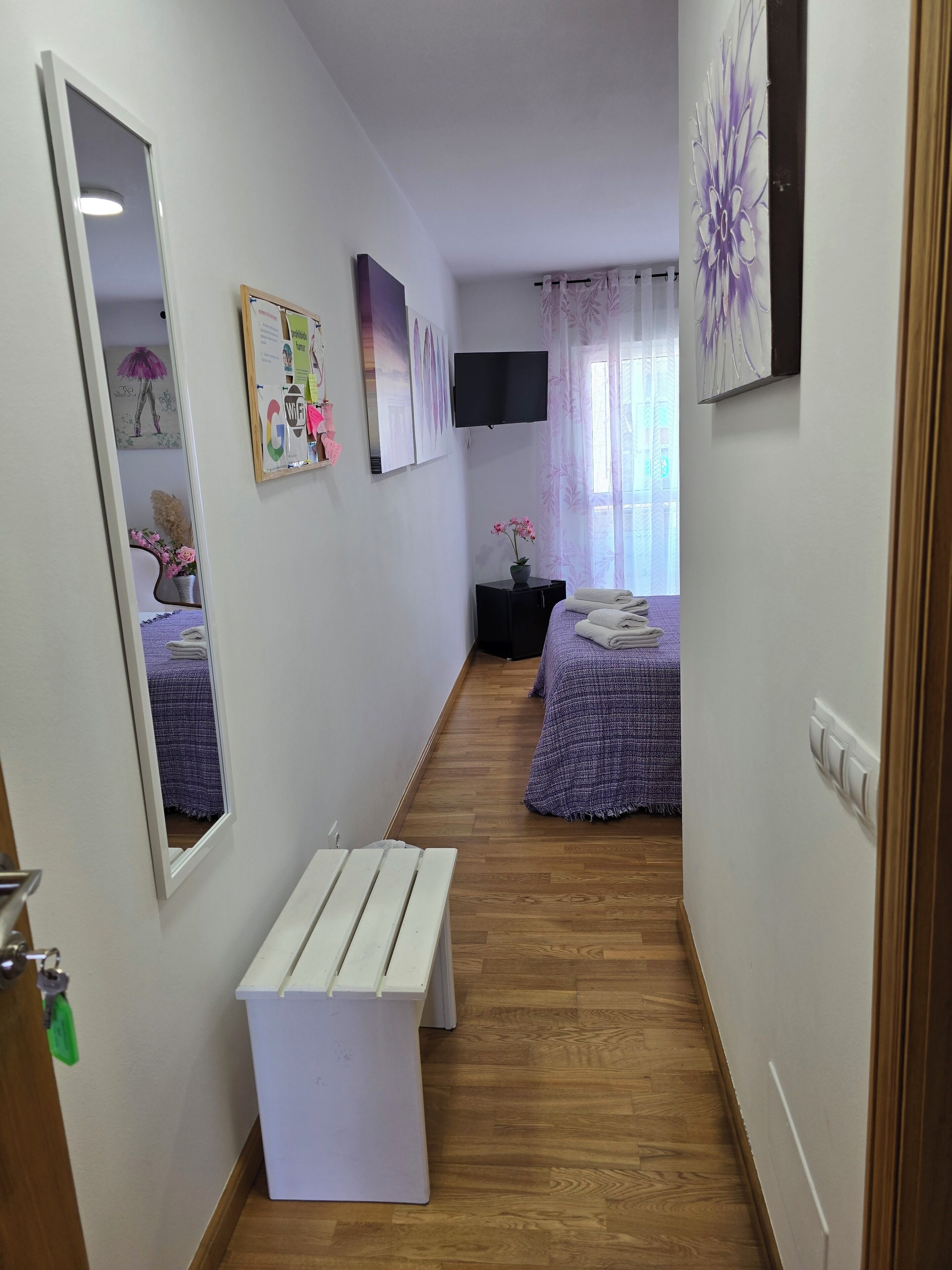A narrow hallway with a wooden floor leading to two bedrooms, each with a purple bedspread and a small bedside table. A mirror and a framed picture are on the left wall, and a TV is mounted above the bed on the right.