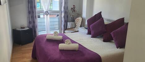 Una cama bien tendida con ropa de cama morada y blanca, una mesita de noche con una lámpara y vistas al exterior a través de una ventana con cortinas.