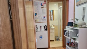 A hallway with a wooden door, a white refrigerator, and a white shelf unit with various items. - Pensión Myrian (O Pino)