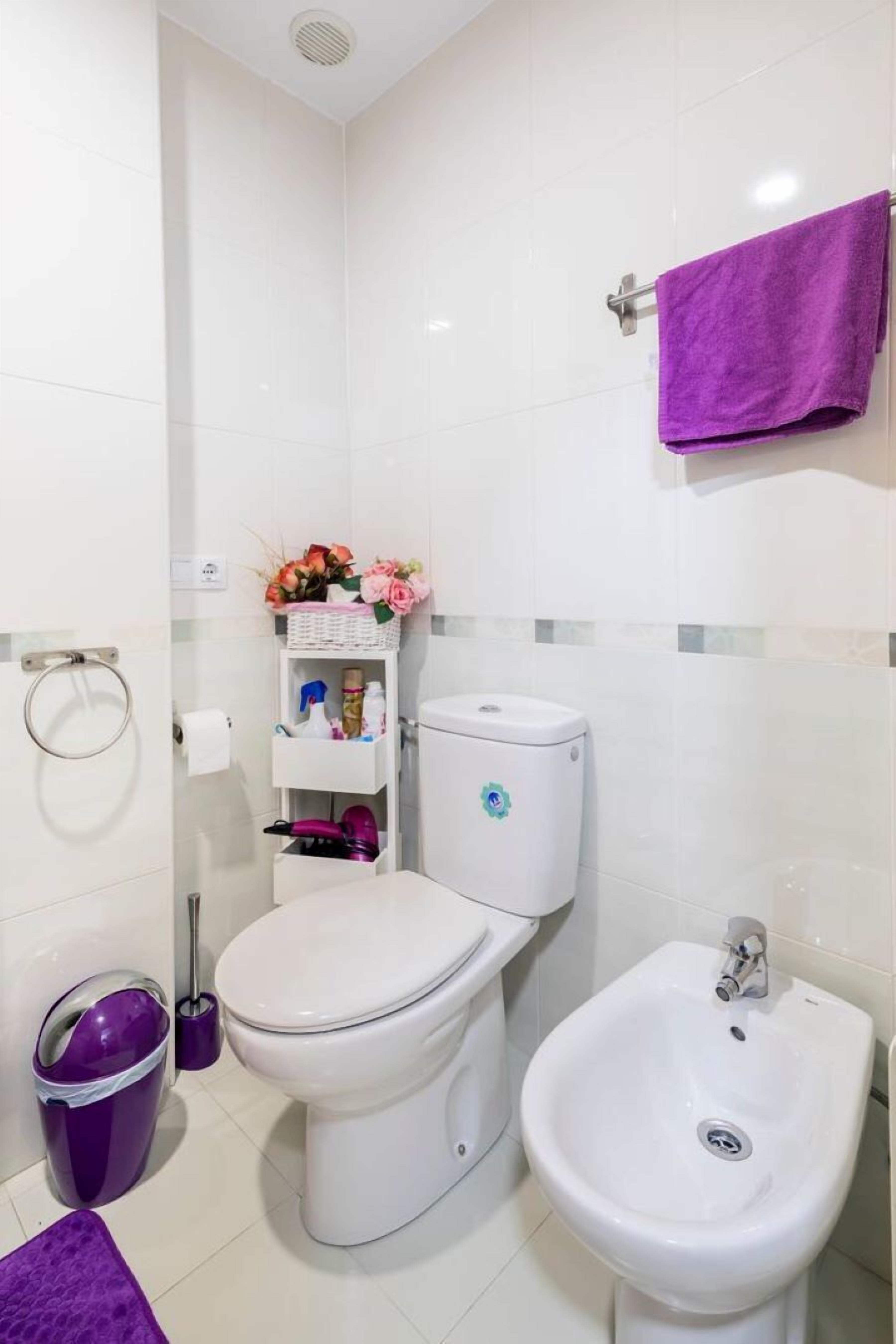 Un baño con inodoro, bidé y un estante con artículos de aseo y un arreglo floral.