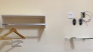 A room with a white air conditioner, a wooden hanger, a white shelf, and a glass shelf with two black objects.