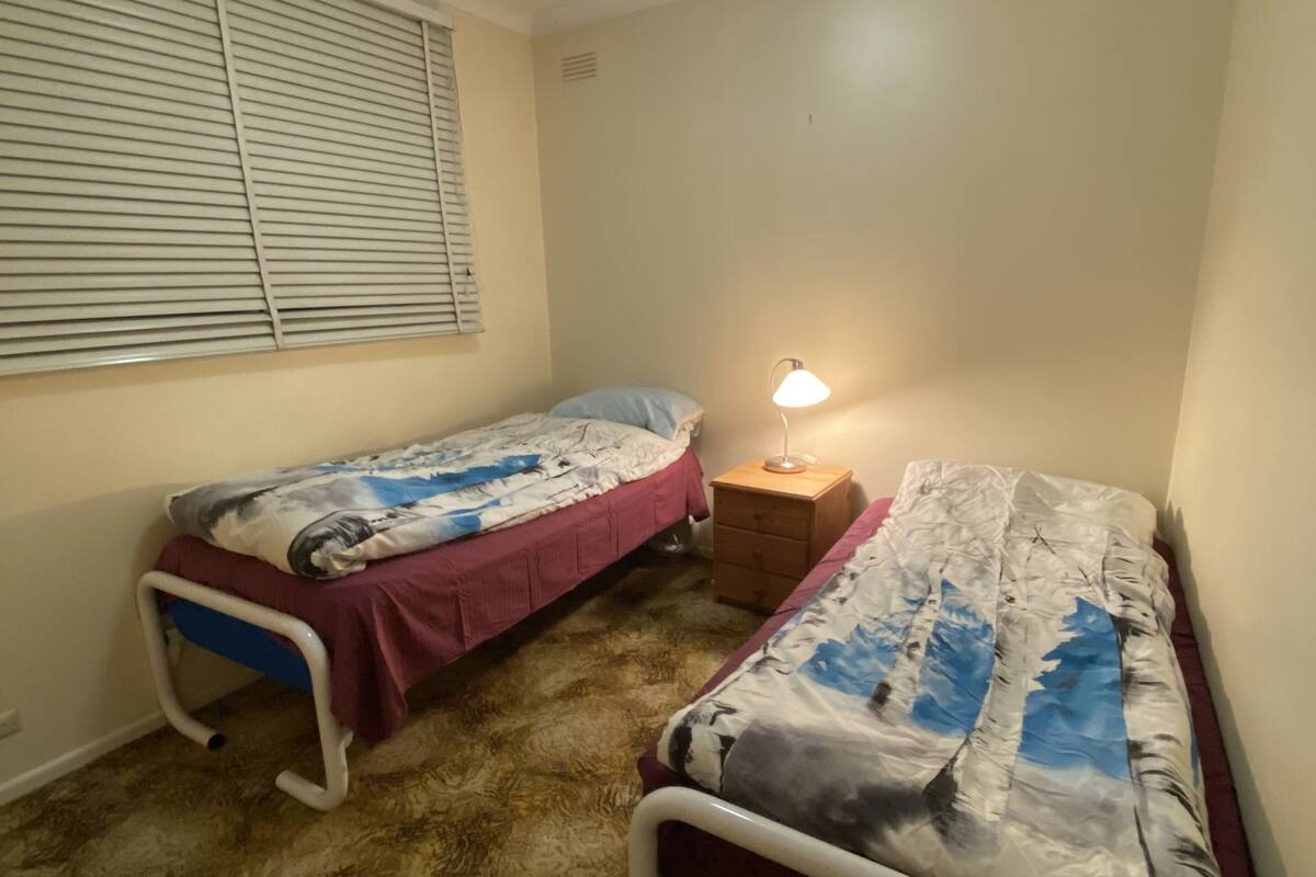 A small room with two single beds, a bedside table with a lamp, and a window with blinds.
