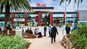 Un centro comercial Westfield con una sucursal del banco HSBC, palmeras y personas caminando y sentadas en el área exterior.