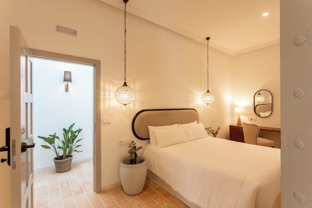 A hotel room with a bed, bedside table, lamp, potted plants, and a door.
