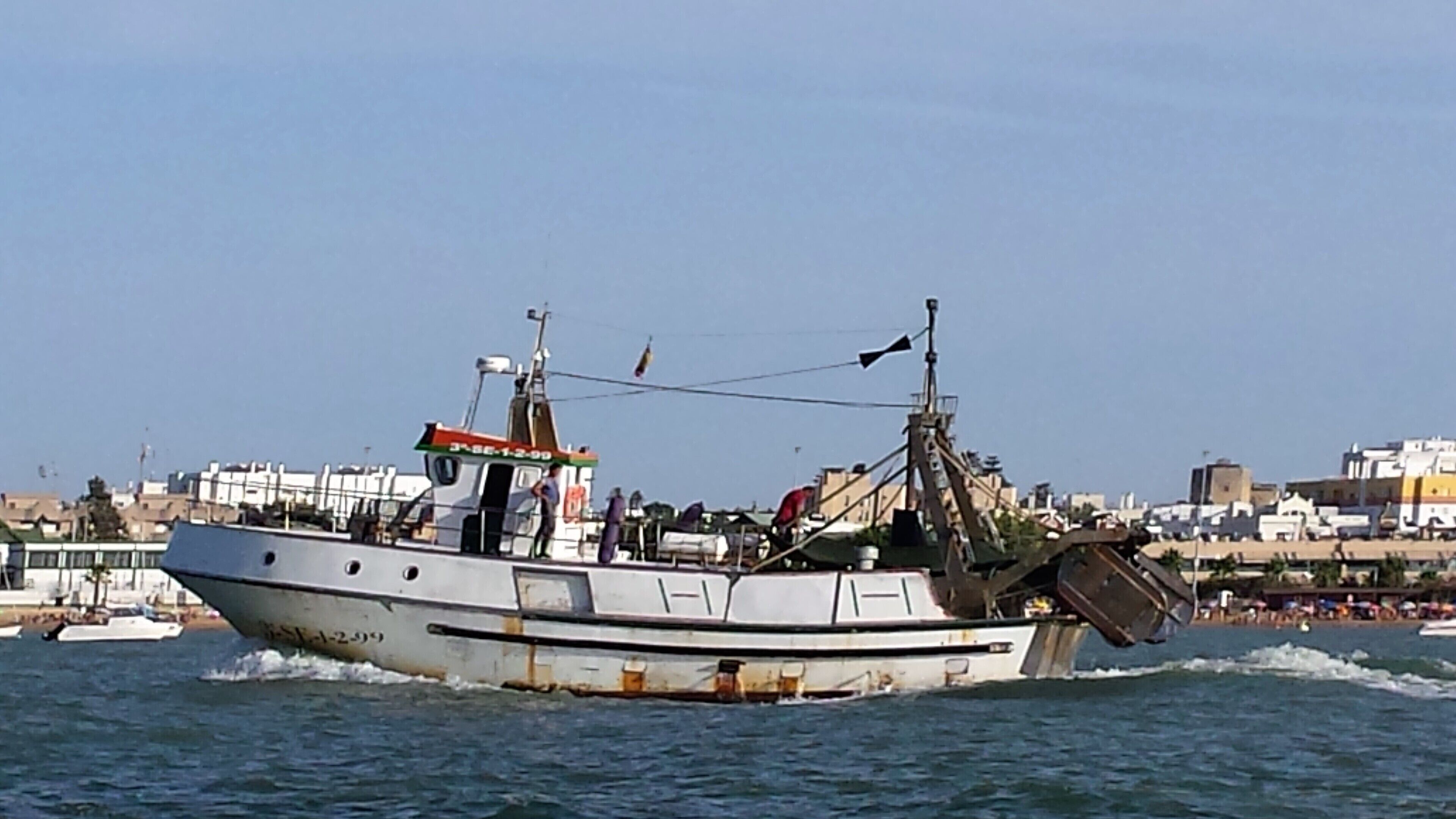 Um barco com cabine e equipamentos de convés navegando em águas, com paisagem urbana ao fundo.