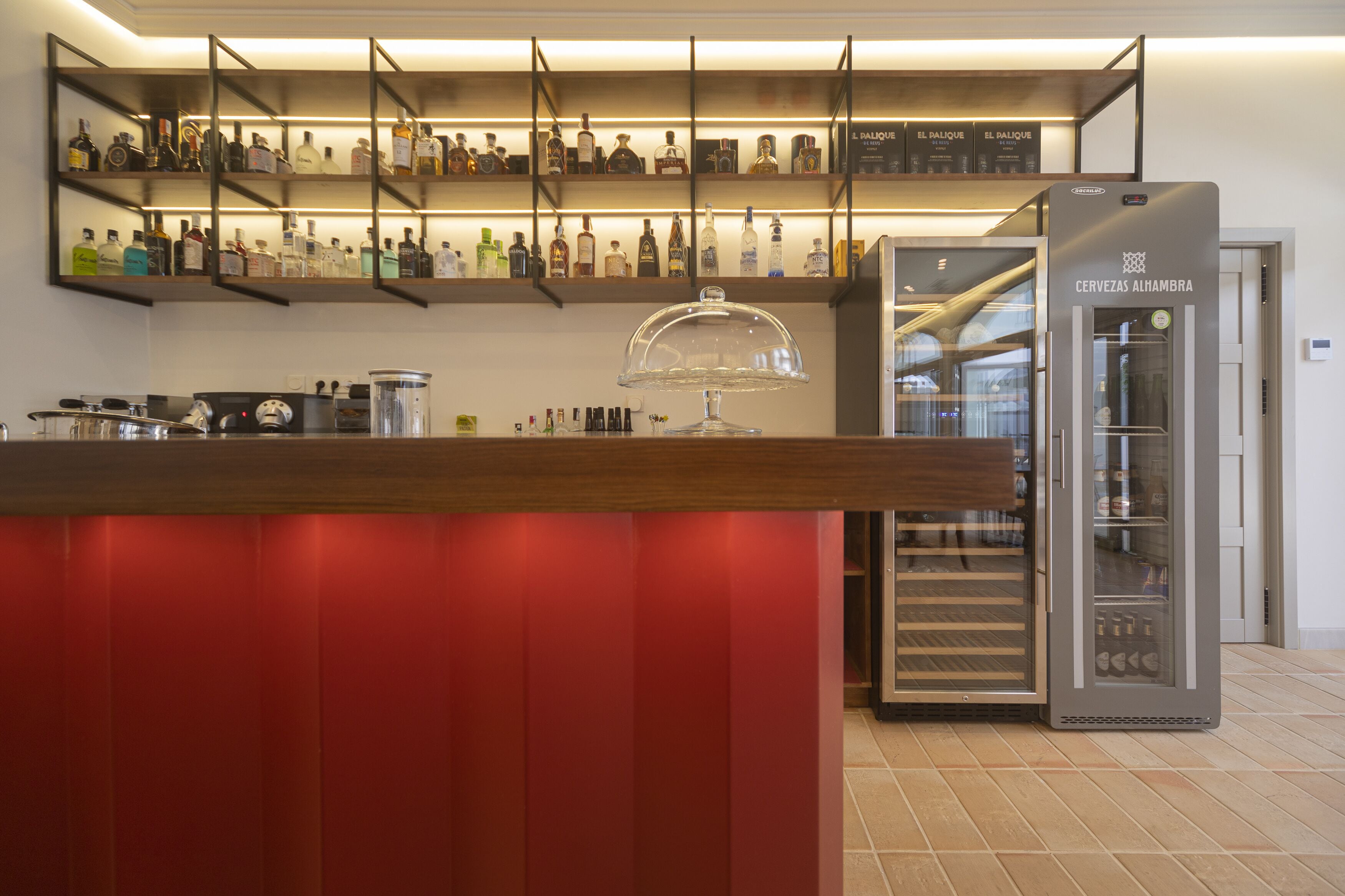 Zona de bar con estantes de botellas de licor, un expositor de vidrio y un refrigerador con la etiqueta 'CERVEZAS ALIMBRA'.