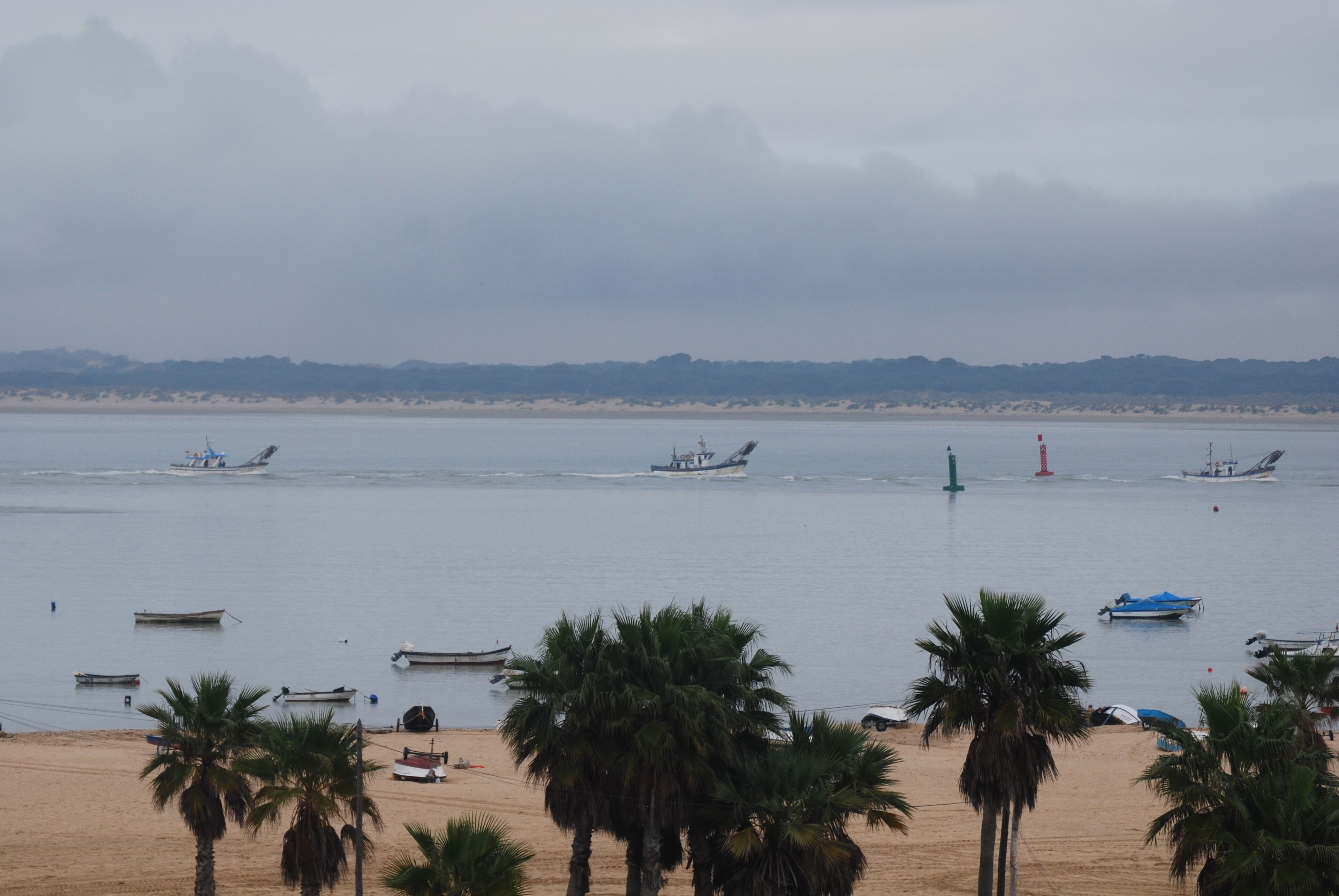 Una playa con barcos, palmeras y un faro a lo lejos.