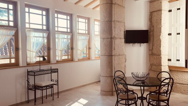 A spacious room with a high ceiling, exposed wooden beams, large windows with sheer curtains, a stone pillar, a round table with chairs, and a flat-screen TV mounted on the wall. - Casa Guardião (Chaves)
