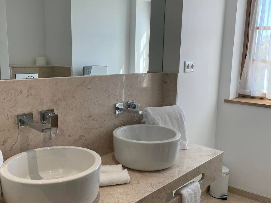 Un baño moderno con dos lavamanos grandes de color blanco, un espejo amplio y una ventana con cortinas.