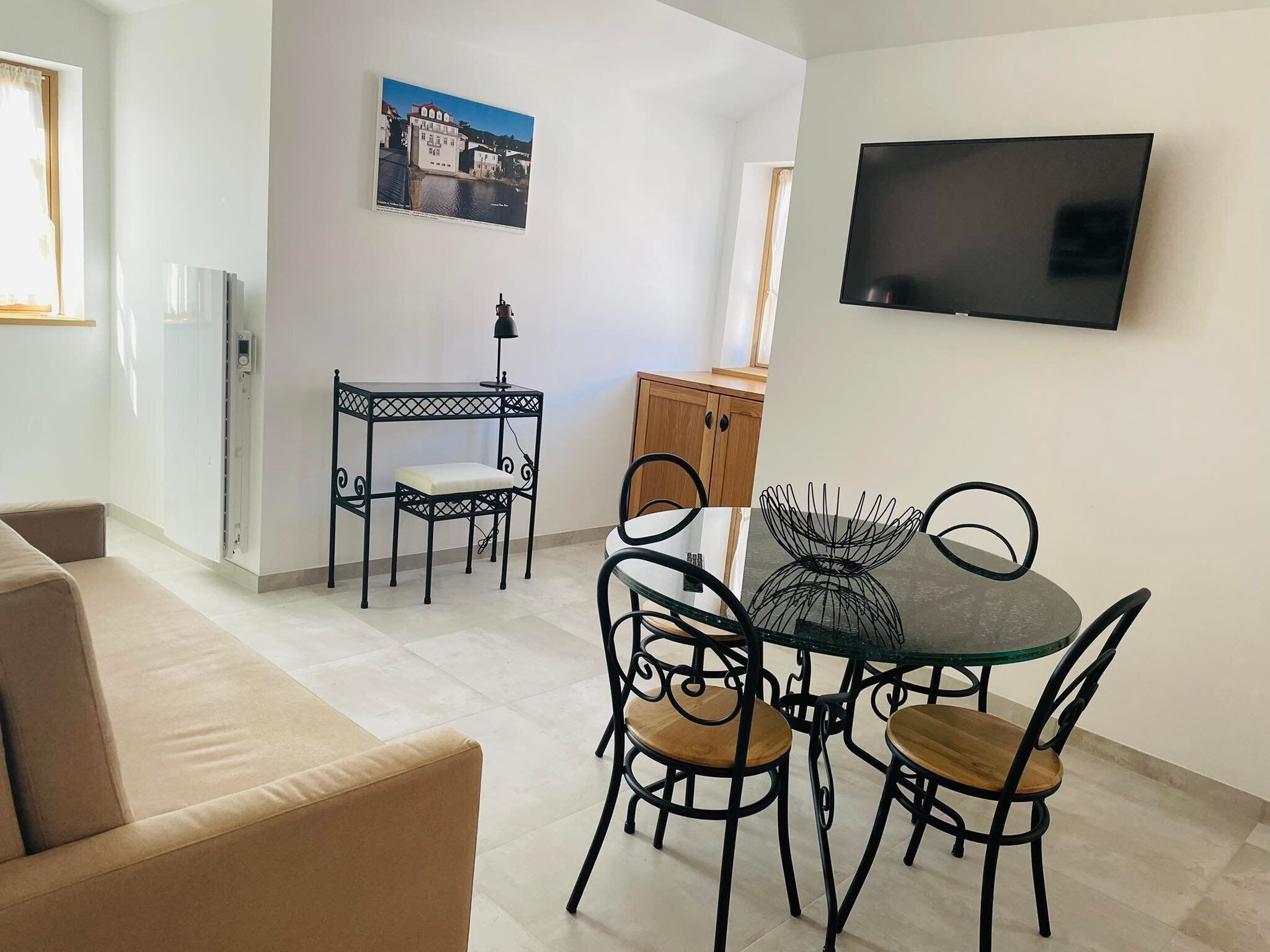 Een woonkamer met een beige bank, een eettafel met vier stoelen, een klein bijzettafeltje met een lamp, en een tv aan de muur.