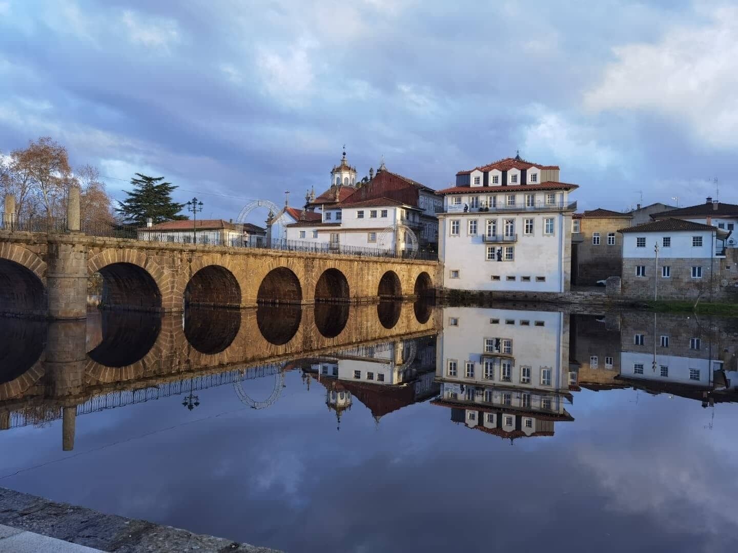 Un puente de piedra con arcos sobre una masa de agua tranquila, que refleja edificios y una noria al fondo.
