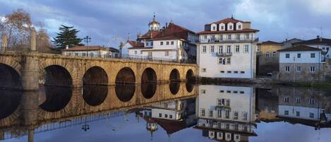 Un puente de piedra con arcos sobre una masa de agua tranquila, que refleja edificios y una noria al fondo.
