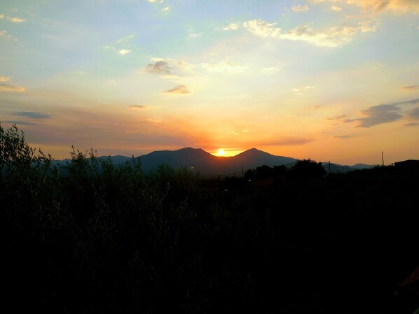 A sunset over a mountain range with silhouettes of vegetation and distant buildings.