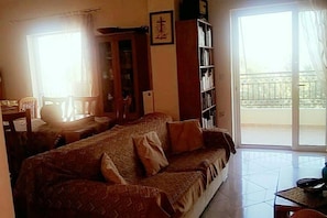 Uma sala de estar com sofá, mesa de jantar com cadeiras, estante de livros e porta de vidro deslizante para uma varanda.