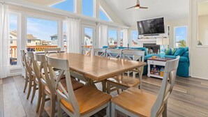 A spacious living area with a large wooden dining table, multiple chairs, a teal sofa, and a flat-screen TV mounted on the wall.