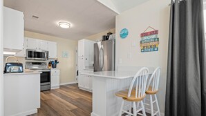 A modern kitchen with white cabinetry, a stainless steel refrigerator, a microwave, and an oven. There are two wooden chairs at a small island.
