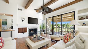A spacious living room with a fireplace, comfortable seating, and a large window offering a view of the outdoors. - Los Estados #66 - 3 BD /3.5 BA, 2586sf (La Quinta)