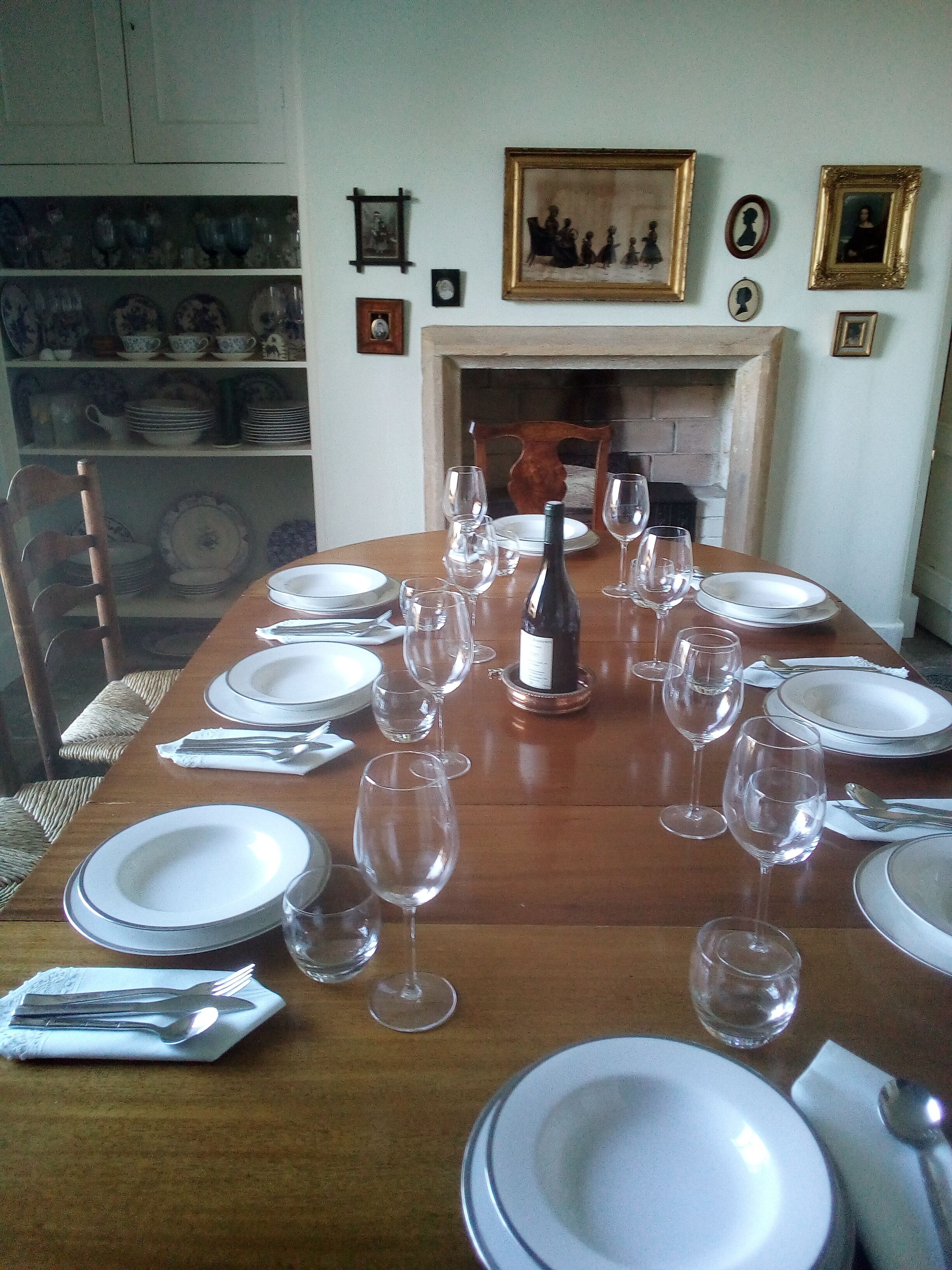 A dining table set for six with plates, glasses, and cutlery. There is a fireplace in the background.