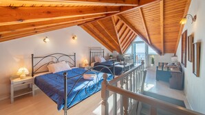A high-ceilinged room with a wooden beam structure, two beds with blue bedding, a small table with a lamp, and a view of the outside through a window. - Holiday Home "Balis Panorama" with Sea View, Balcony, Garden & Wi-Fi (Corfu)