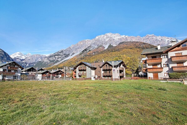 Une station de montagne avec des bâtiments de style chalet, un espace verdoyant au premier plan et des montagnes enneigées en arrière-plan.