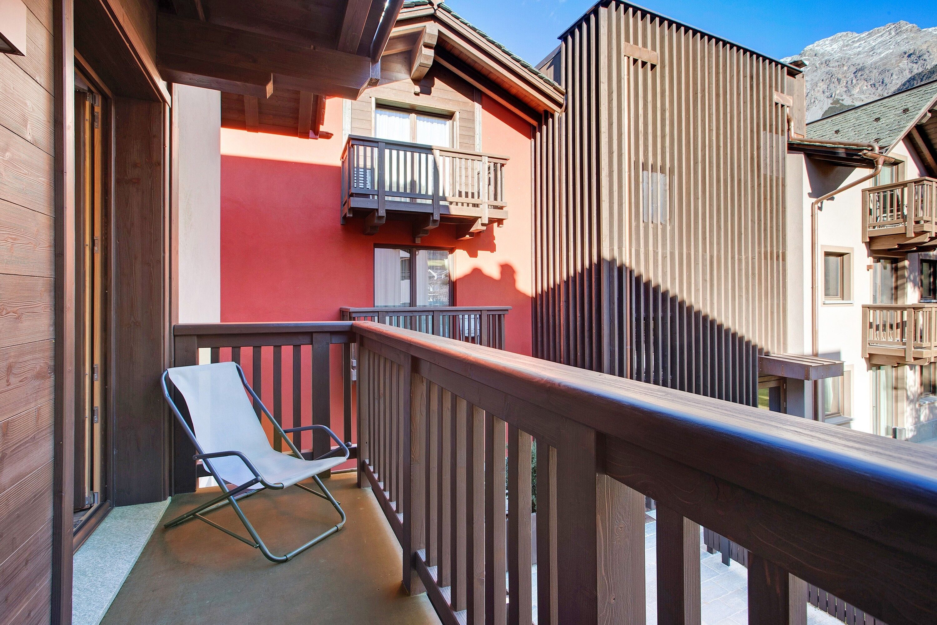 A balcony with a wooden railing, a white chair, and a building with a red facade and balconies.