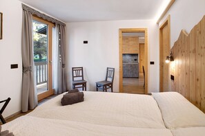 A bedroom with a large bed, two chairs, a wooden door, and a view of a kitchen through an open door.