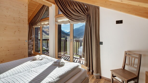 A bedroom with a bed, a wooden chair, a window with curtains, and a balcony with a view of mountains. - Apartment "Baita Emilia e Daniele 13" with Mountain View, Balconies and Wi-Fi (Bormio)