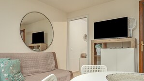 Una habitación con un espejo redondo, un televisor de pantalla plana, un sofá con cojines estampados, una mesa de comedor con sillas y un mueble con un televisor encima.