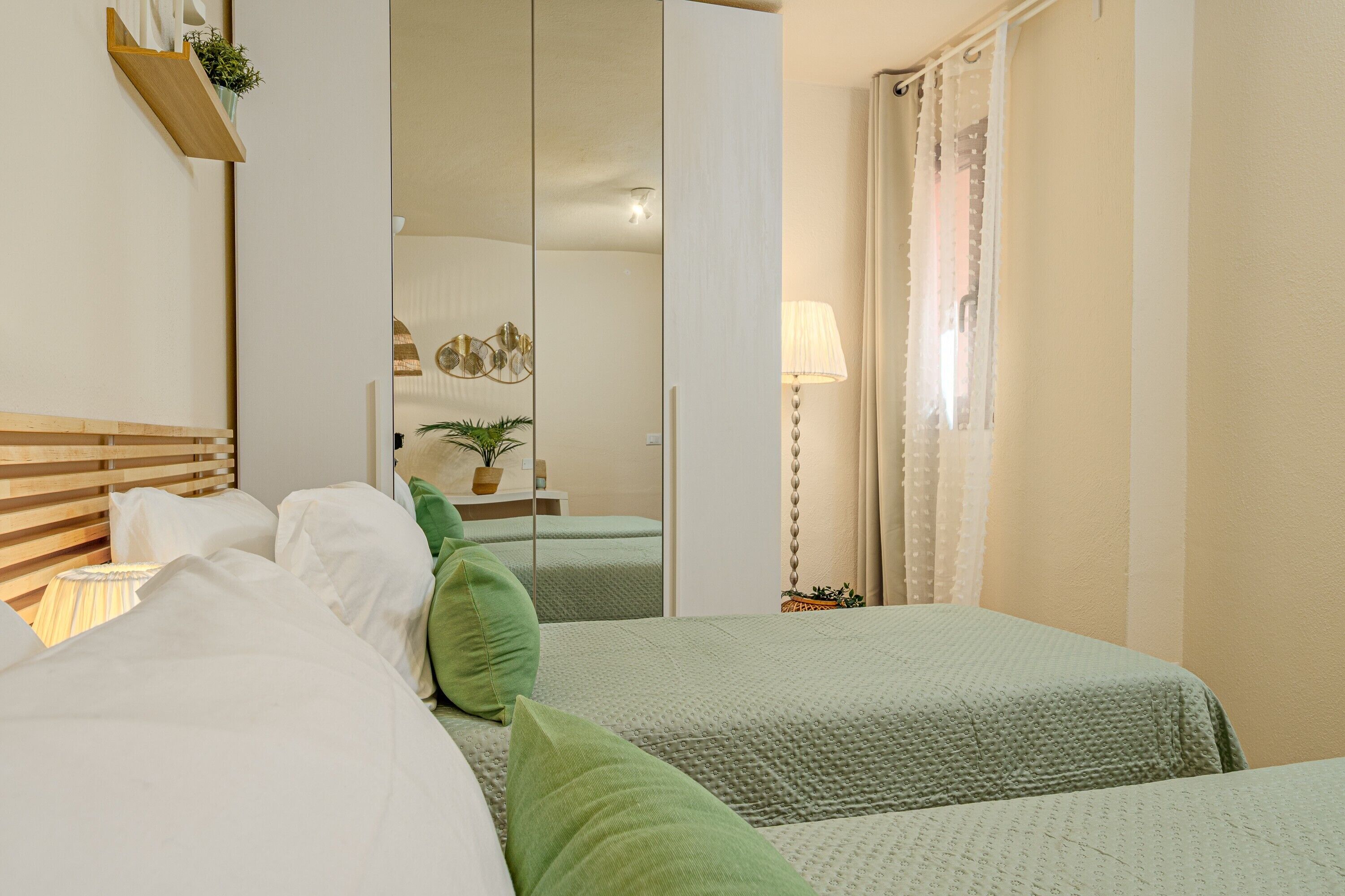 A hotel room with two beds, a mirror, a bedside table with a lamp, and a window with curtains.