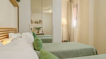 A hotel room with two beds, a mirror, a bedside table with a lamp, and a window with curtains.