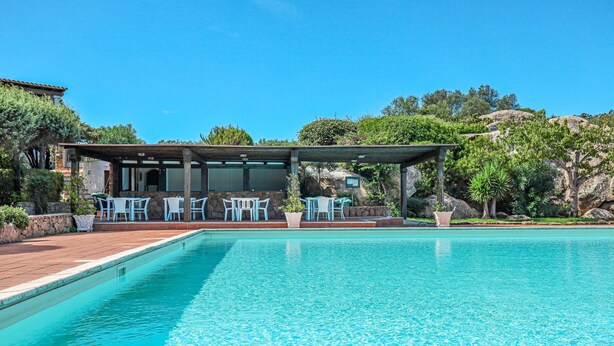 Una piscina con un porche cubierto, rodeada de vegetación y un cielo azul despejado.