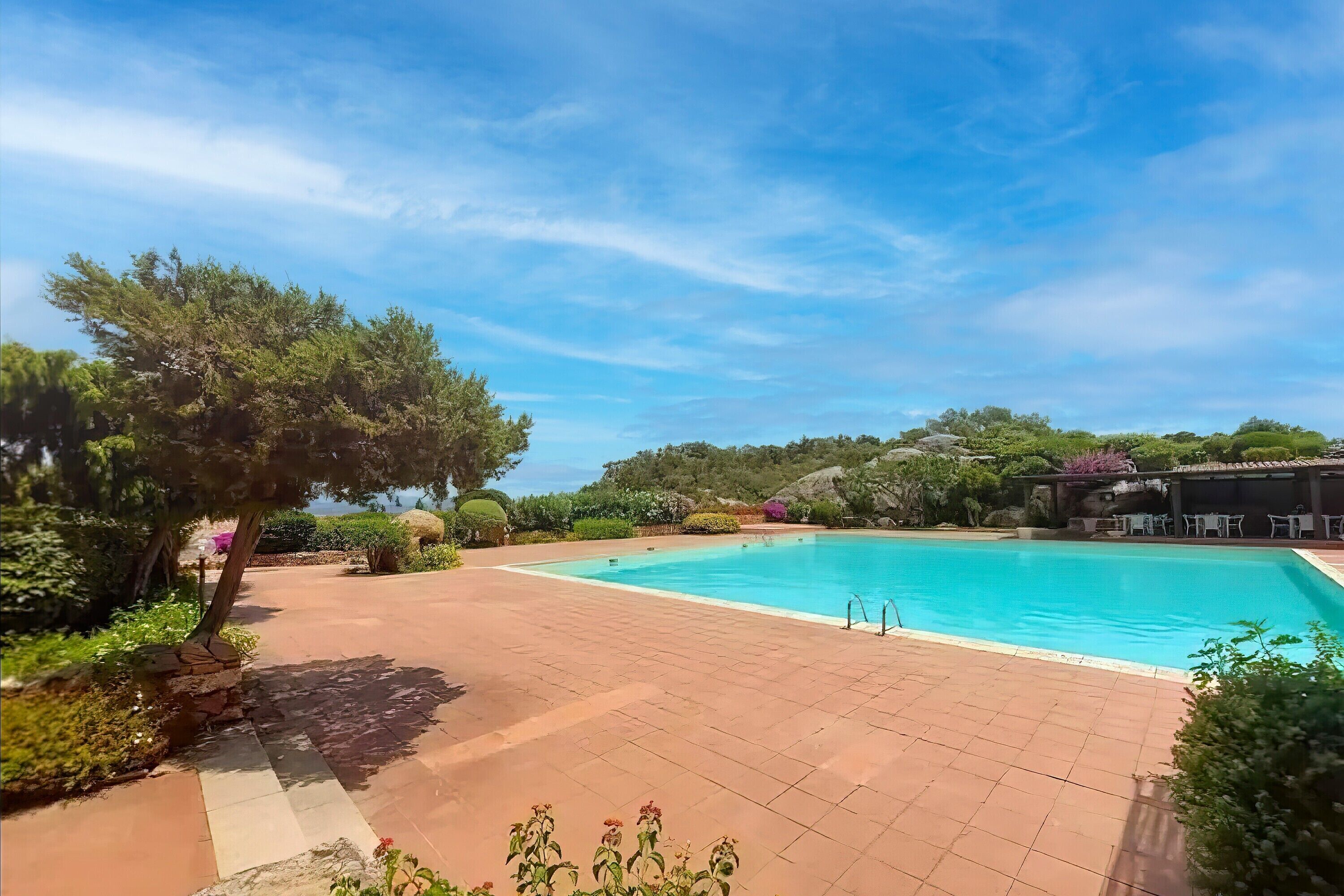 Une piscine dotée d’une terrasse carrelée, entourée de verdure et surplombée par un ciel dégagé.