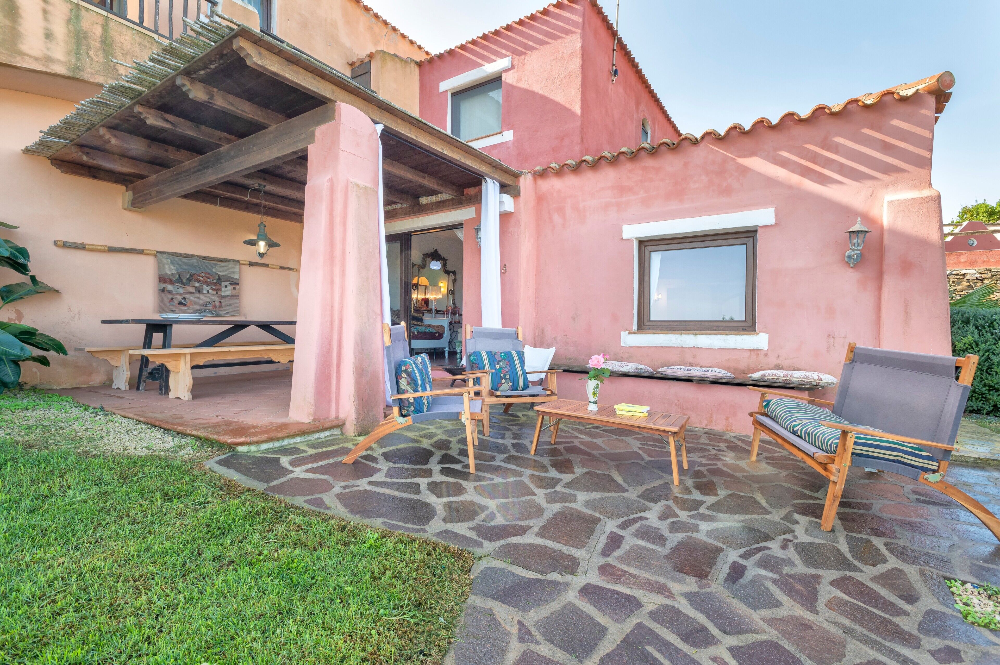 A patio with stone flooring, wooden furniture, and a view of a pink building with a window and a door.