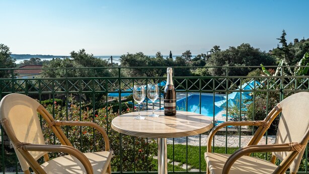 A balcony with wicker chairs, a table with a bottle and glasses, and a pool in the background. - Studio Apartment "Anemona 3" with Shared Pool & Wi-Fi (kefalonia)