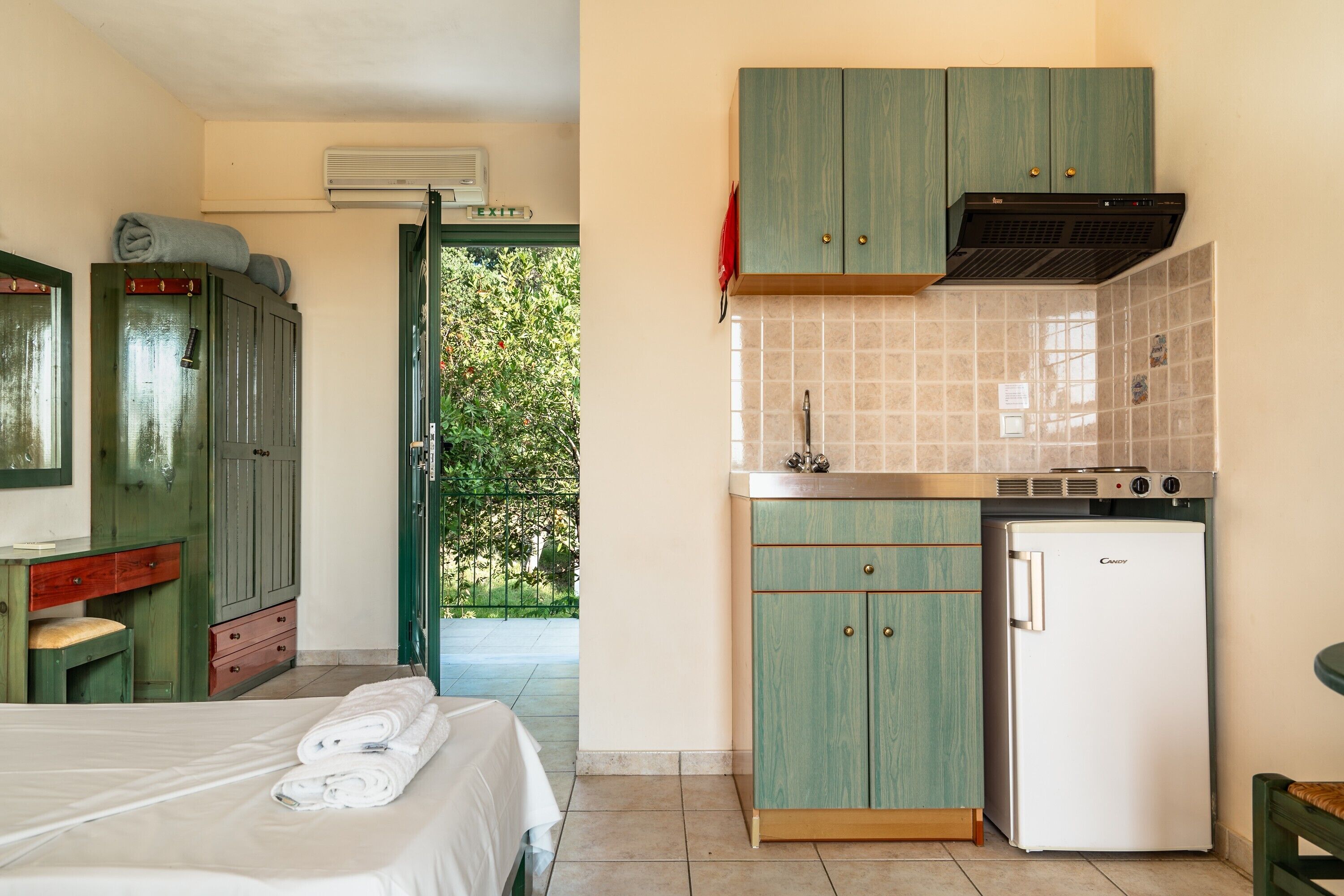 A room with a bed, a kitchenette, and a view of greenery outside.