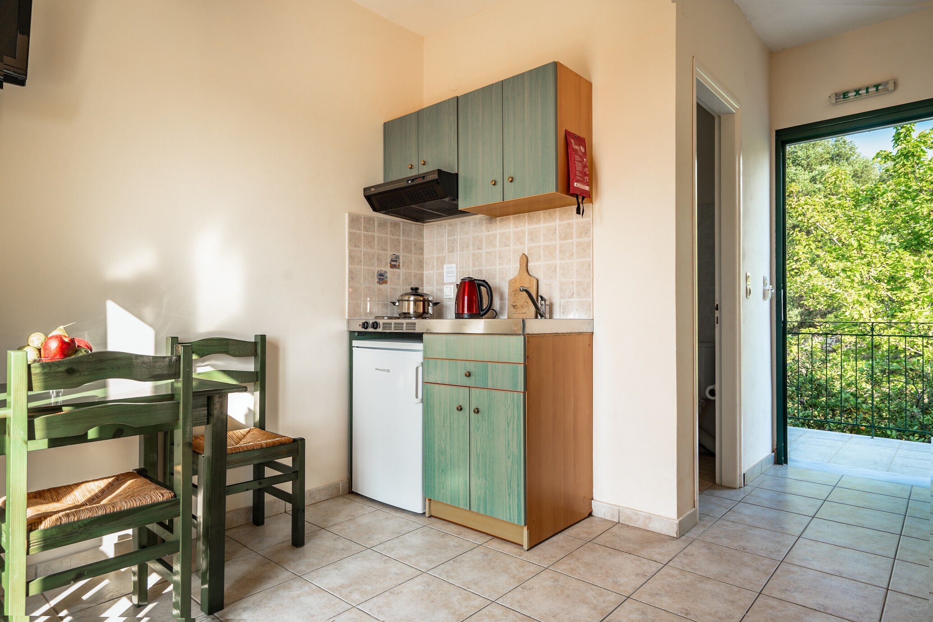 A compact kitchen with a built-in dishwasher, a wooden chair, and a view through an open door to a balcony.