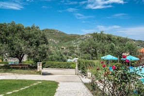 A paved pathway leading to a bench and a stone wall, surrounded by lush greenery and a mountainous backdrop.
