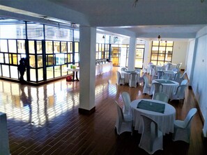 A spacious indoor area with multiple round tables covered with white tablecloths and chairs, large windows with blinds, and a tiled floor. - Virunga Hotel (Muhoza)