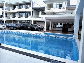 A swimming pool with a tiled deck, surrounded by a multi-story building with balconies and a parking area with cars. - Virunga Hotel (Muhoza)