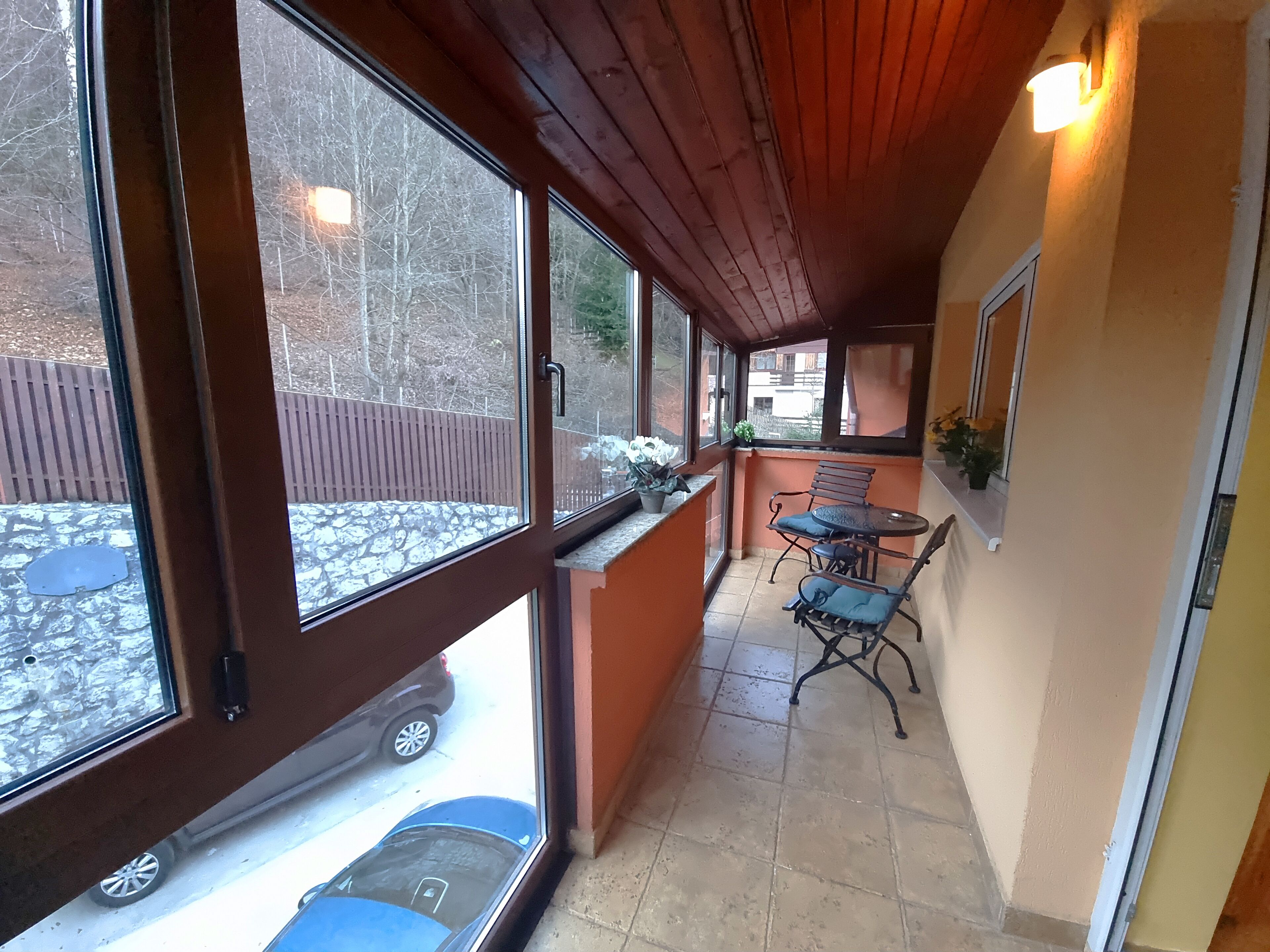 Een overdekt buitengebied met een houten plafond, een kleine tafel met stoelen, en uitzicht op een geparkeerde auto en bomen.