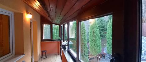 A covered outdoor area with a bench, a small stool, and a view of a garden with trees and a wooden deck.