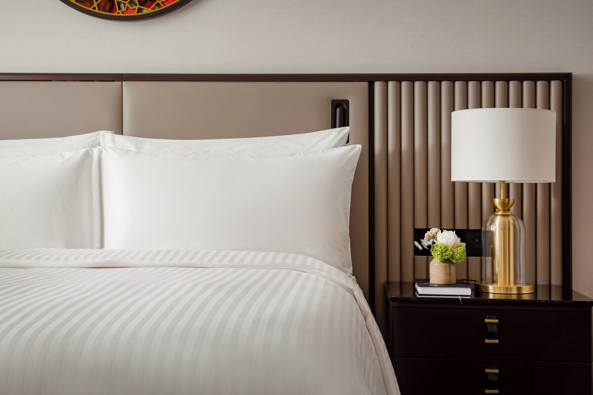 A neatly made bed with white linens, a bedside table with a lamp, and a decorative vase with flowers.