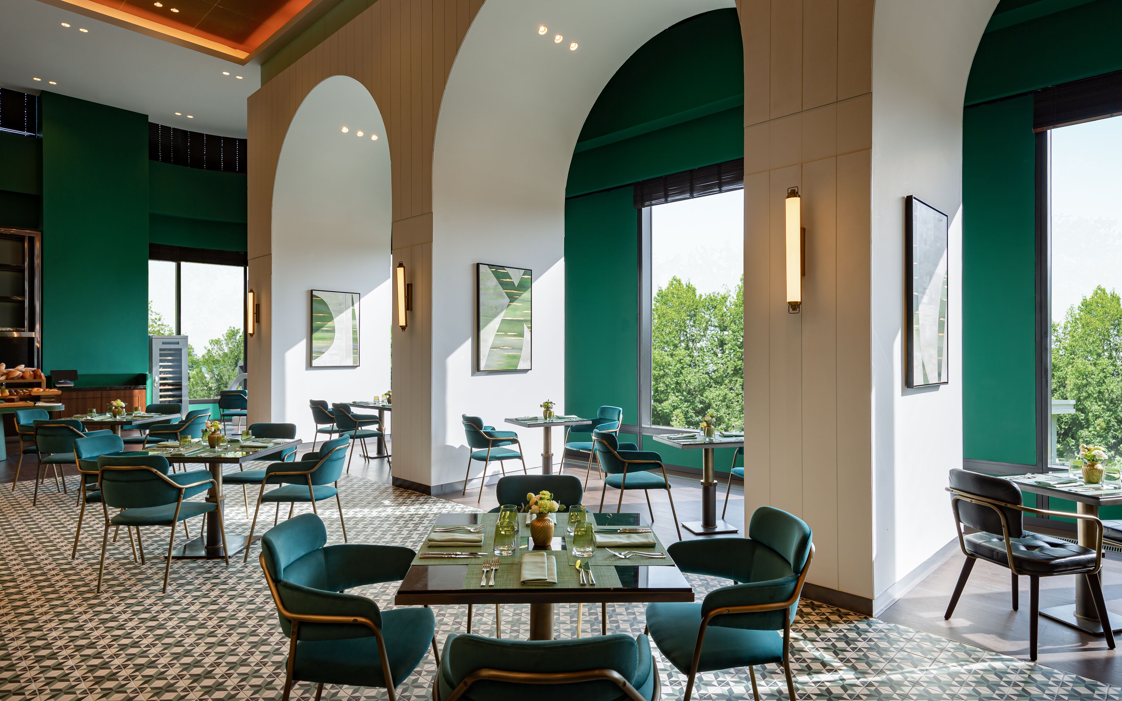 Un comedor amplio con techos altos, paredes verdes y ventanas grandes que ofrecen vista a árboles. Las mesas están dispuestas con sillas y centros de mesa.