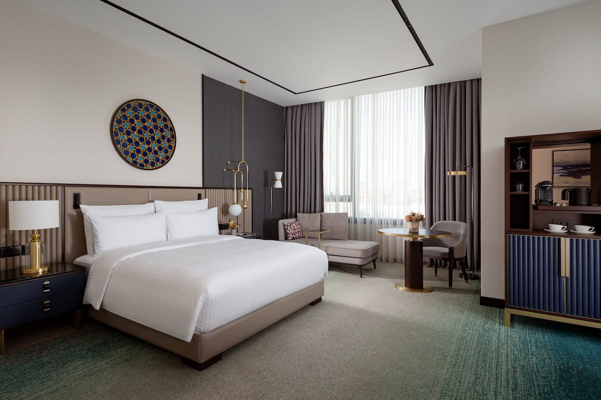 A modern hotel room with a large bed, a desk, a chair, a sofa, and a minibar.