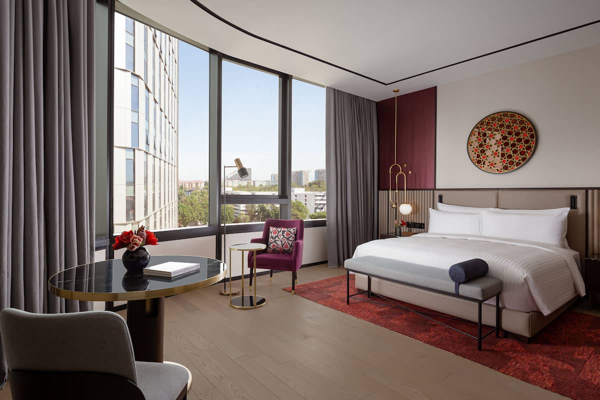 A hotel room with a large bed, a small table, a chair, and a view of the city through large windows.